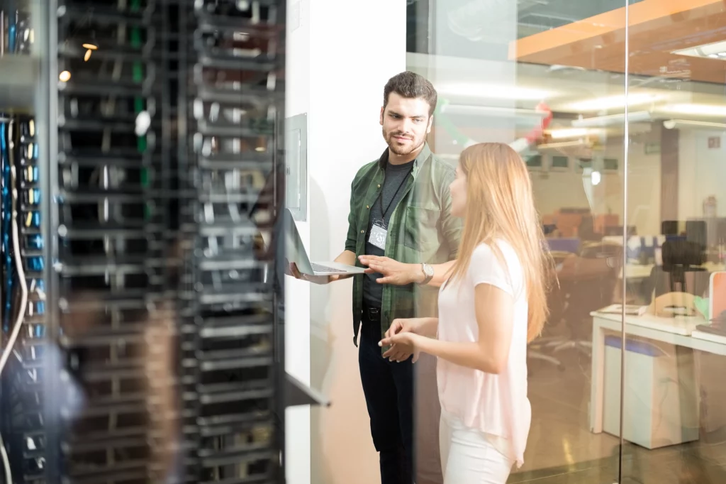 Two people in a server room, one holding a laptop and the other gesturing, discussing technology and data services.