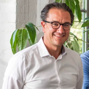 Sam Bambust, CEO of De Cronos Group, wearing glasses and a white shirt, smiles.